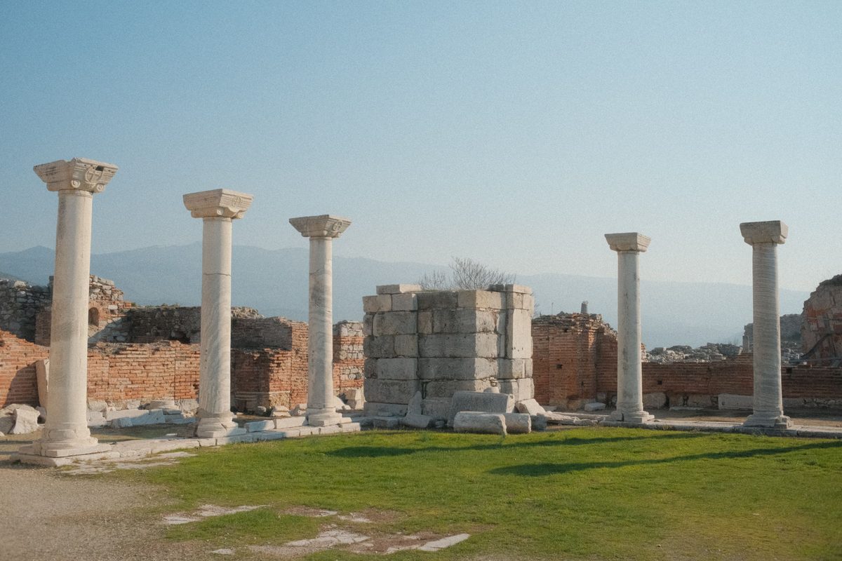 White marble columns with carved capitals standing in the ruined nave of the Basilica of St John on Ayasuluk Hill, with brick walls and mountains behind