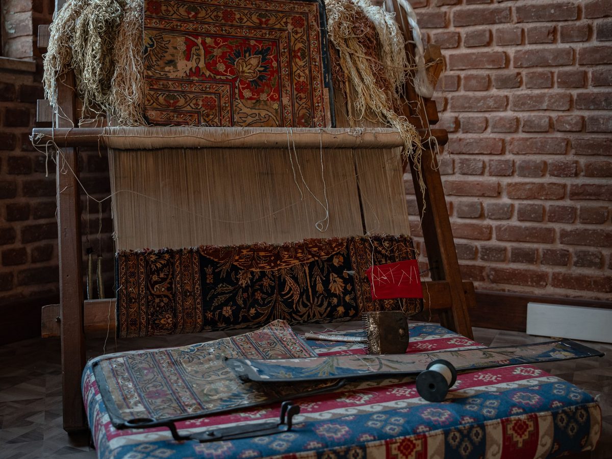 A carpet loom in a brick-walled workshop, with a band of knotted pile at the bottom, bare warp threads above, a finished carpet hung on top and a weaver’s beating comb on the bench
