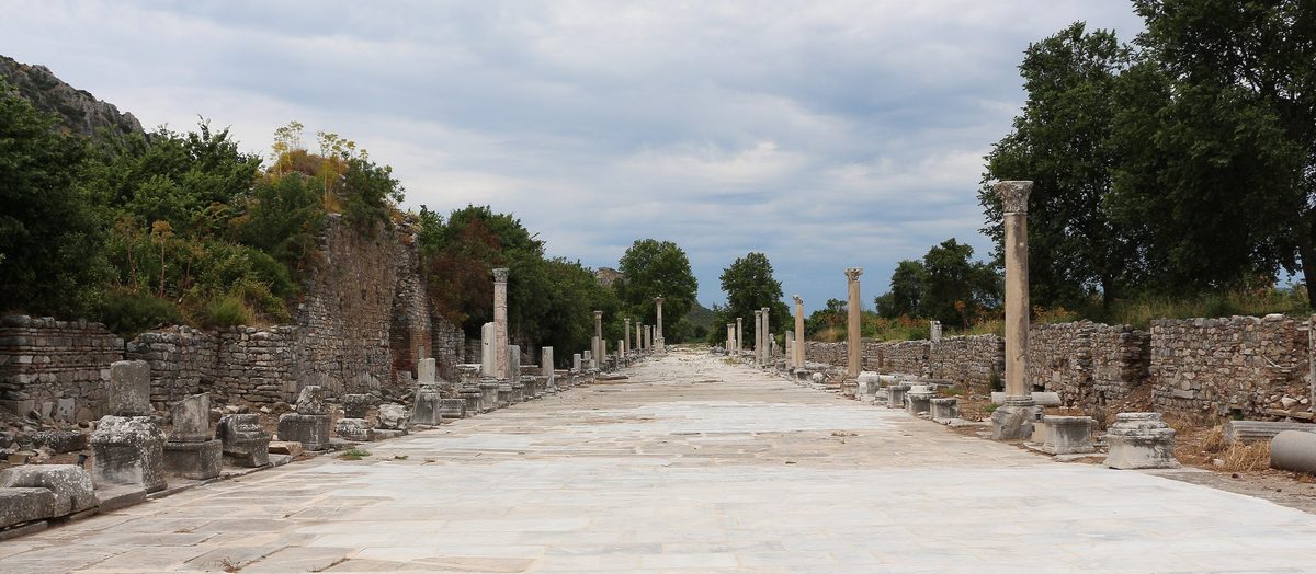 Two rows of standing columns line the wide marble paving of Harbor Street at Ephesus, running away towards wooded ground