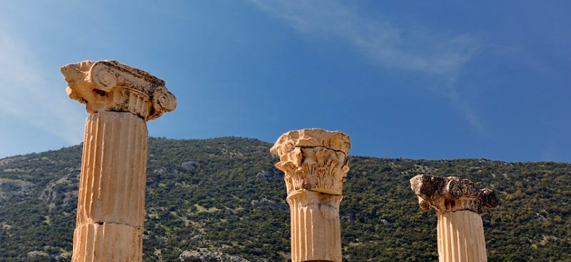 Three ancient column capitals, one Ionic and two Corinthian, rising against a deep blue sky above a green hillside at Ephesus