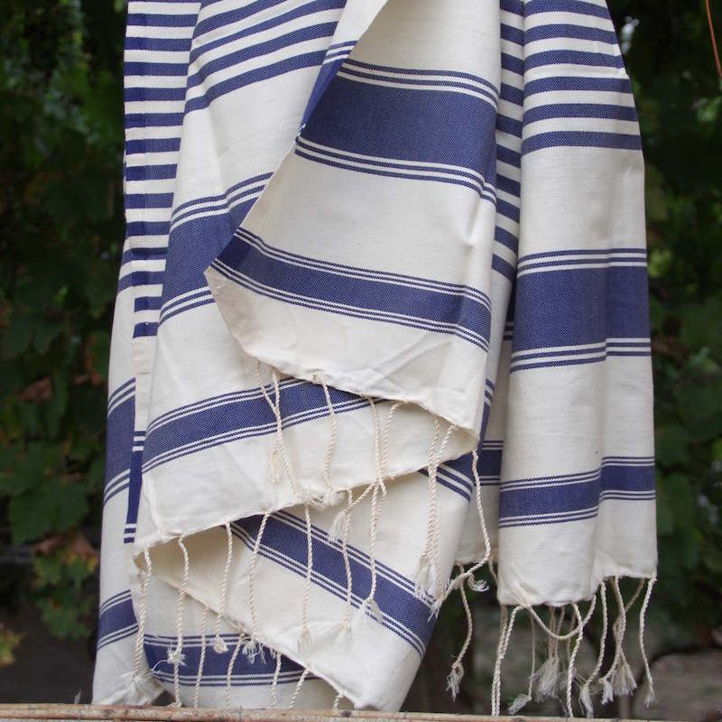 A cream cotton peştemal with navy stripes and knotted fringes hanging outdoors in front of green leaves