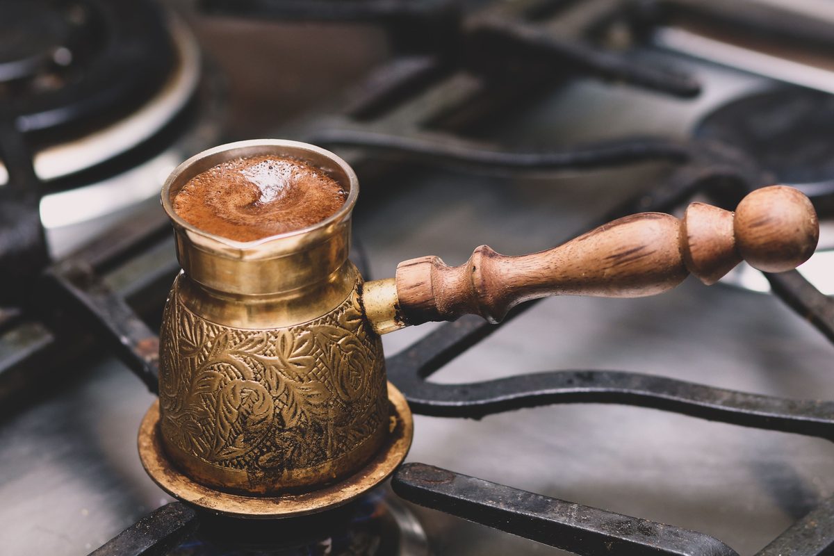 A hand-engraved brass cezve with a turned wooden handle, full of foaming Turkish coffee on a gas stove