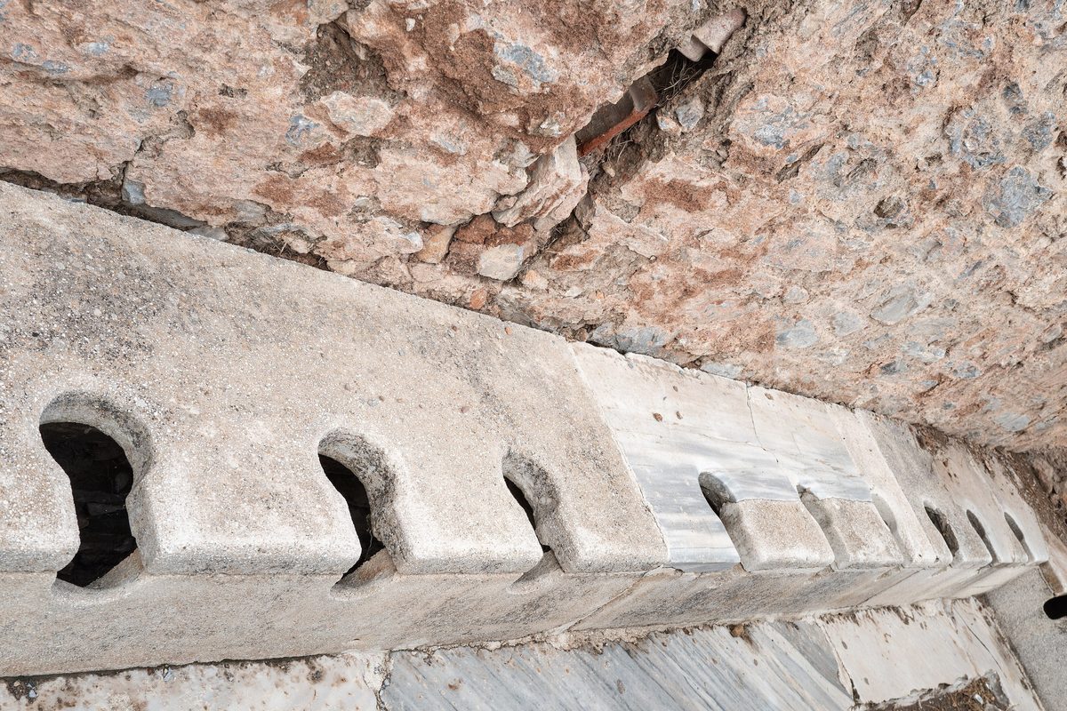 A row of keyhole-shaped openings cut into a long marble bench in the Roman public latrines of Ephesus, against a rough stone wall