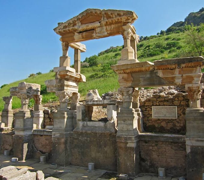 Arched facade and columns of the Roman Fountain of Trajan at Ephesus against a blue sky and green hillside