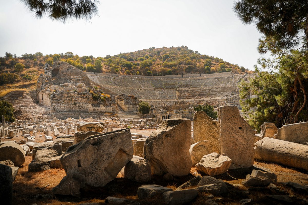 The curved tiers of the Great Theater of Ephesus cut into a green hillside, seen past pine branches and fallen marble blocks in the foreground
