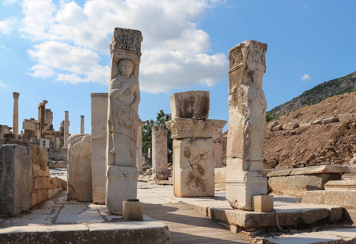 Two tall marble pillars of the Hercules Gate at Ephesus, the left one carved with Hercules wearing a lion skin, with a wooden walkway between them and Curetes Street beyond