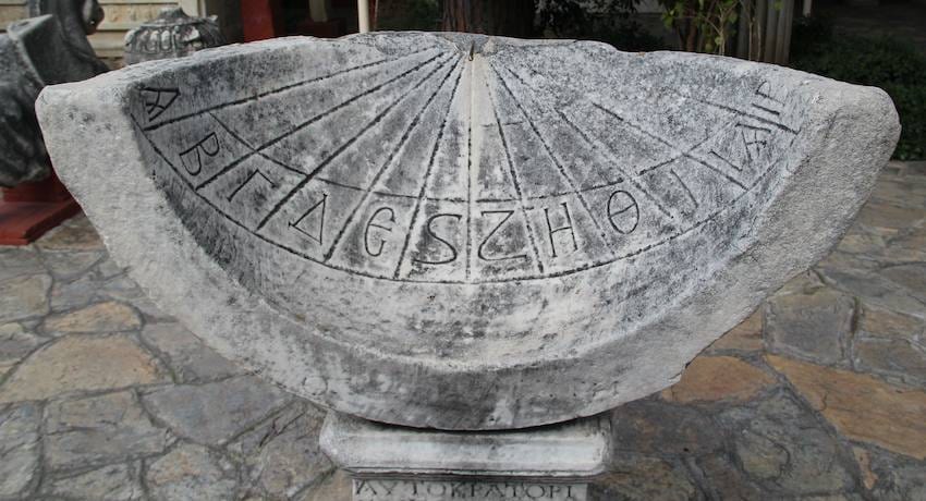 Semicircular ancient marble sundial carved with Greek letters and hour lines, displayed outdoors on a stone plinth