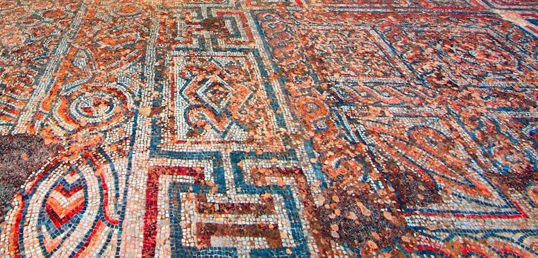 Ancient geometric floor mosaic in red, cream and blue with meander and chevron patterns, laid in small stones