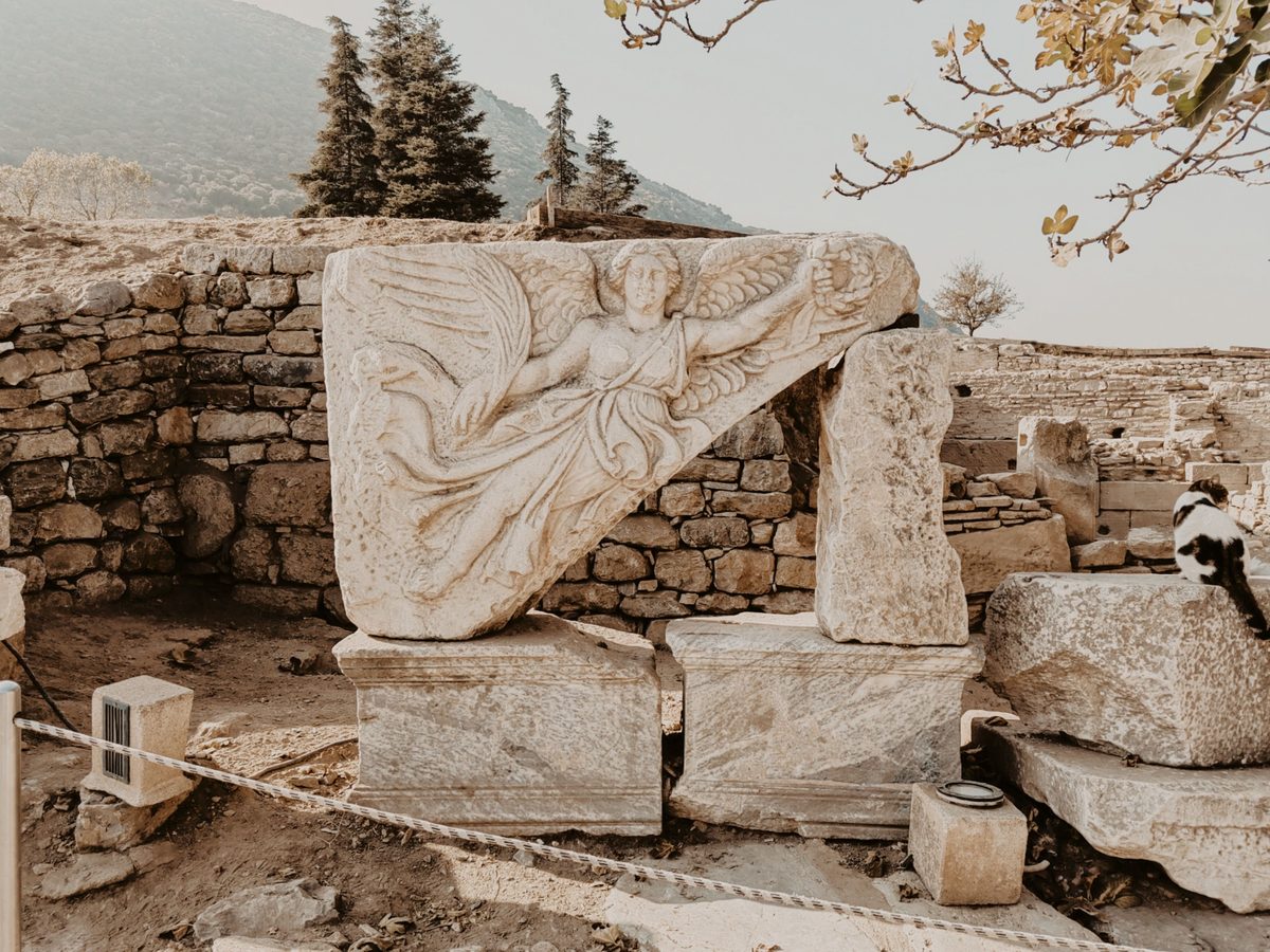 The marble relief of the winged goddess Nike holding out a laurel wreath, set on stone blocks at Ephesus, with a black and white cat sitting on a block to the right