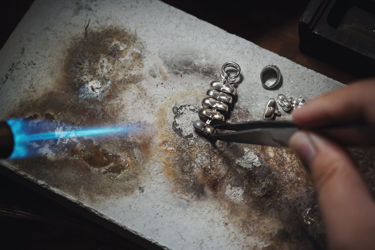 A jeweler’s hand holding small silver pieces with tweezers on a soldering block, a blue torch flame beside them