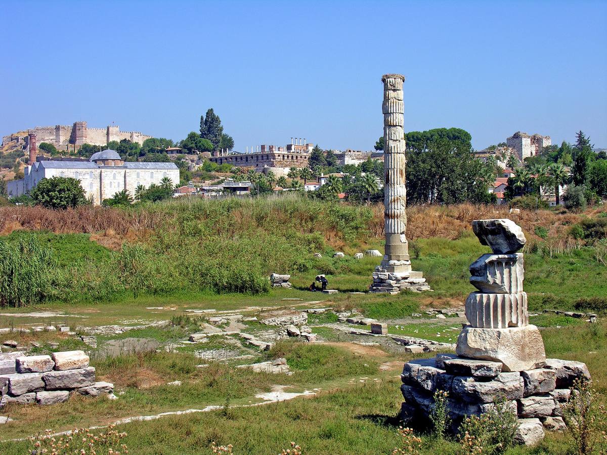 The single re-erected column of the Temple of Artemis in a grassy field, with the İsa Bey Mosque, the Basilica of St John and the fortress walls of Ayasuluk Hill behind it