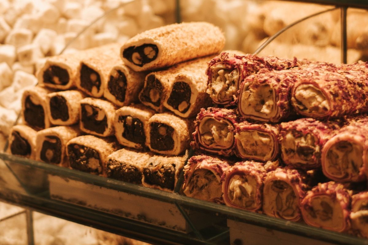 Rolls of Turkish delight filled with nuts, some coated in pale crumbs and some in red flakes, stacked on a glass shelf in front of white sugar-dusted cubes
